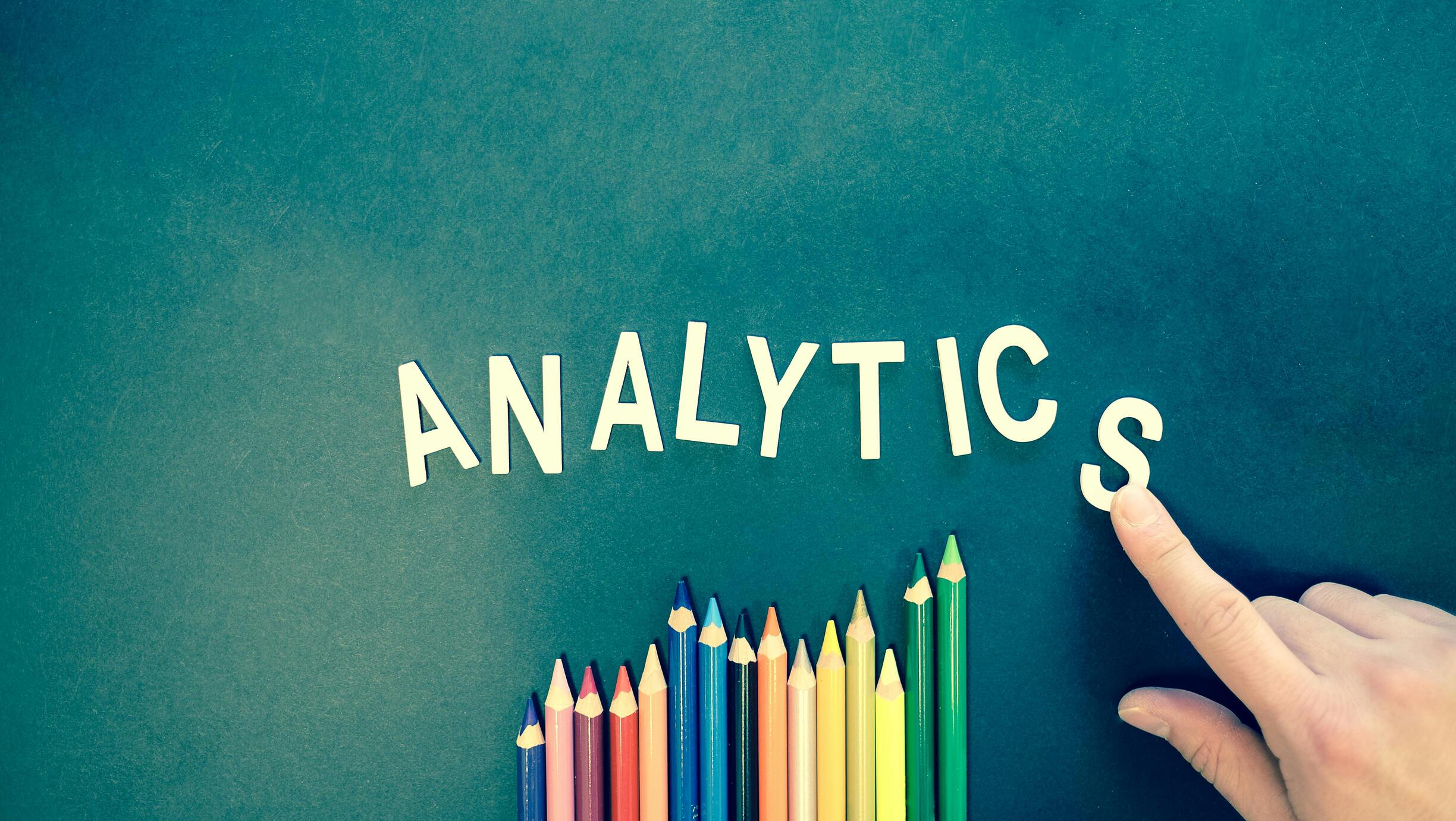 The word “ANALYTICS” in white letters above a row of rainbow-colored pencils arranged like a bar chart